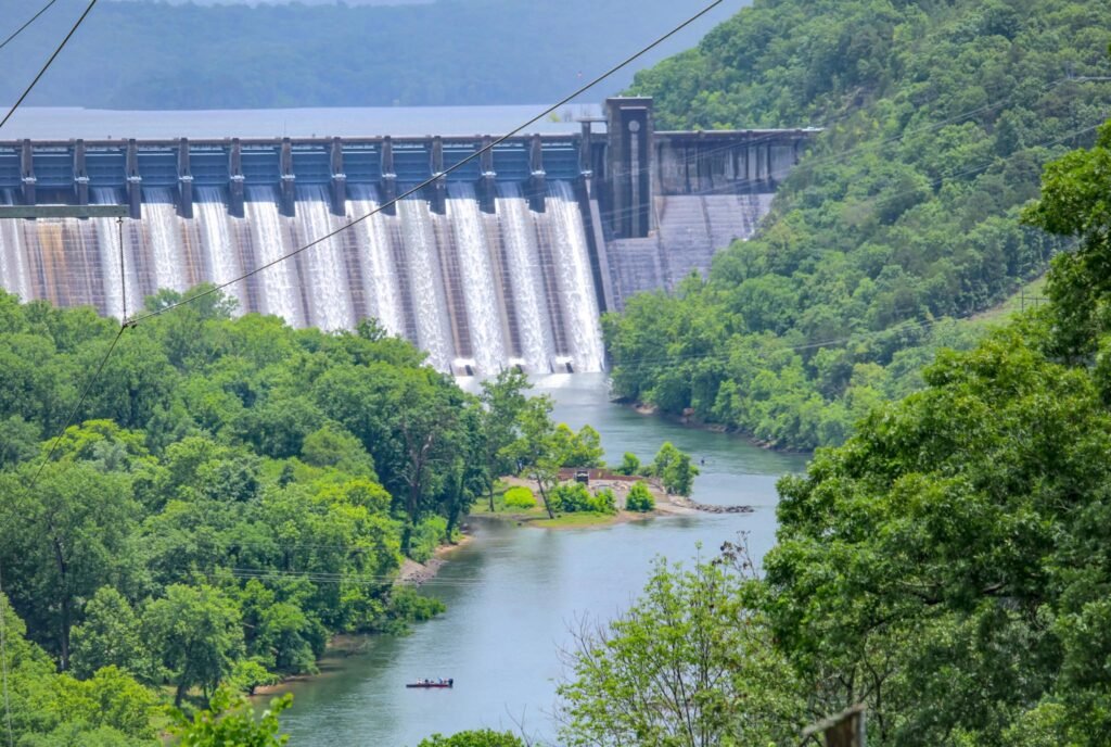 A black-and-white vintage postcard of Norfork Dam showing water cascading down the spillways into the river below, with the lake visible behind the dam and "NORFORK DAM" printed in the bottom right corner.